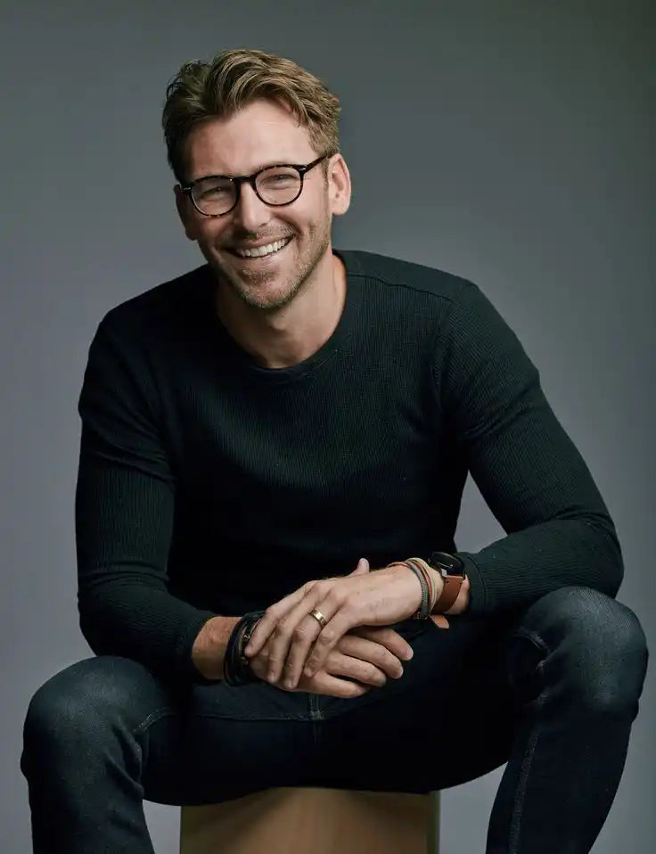 Smiling man with glasses wearing a dark sweater and sitting on a stool.